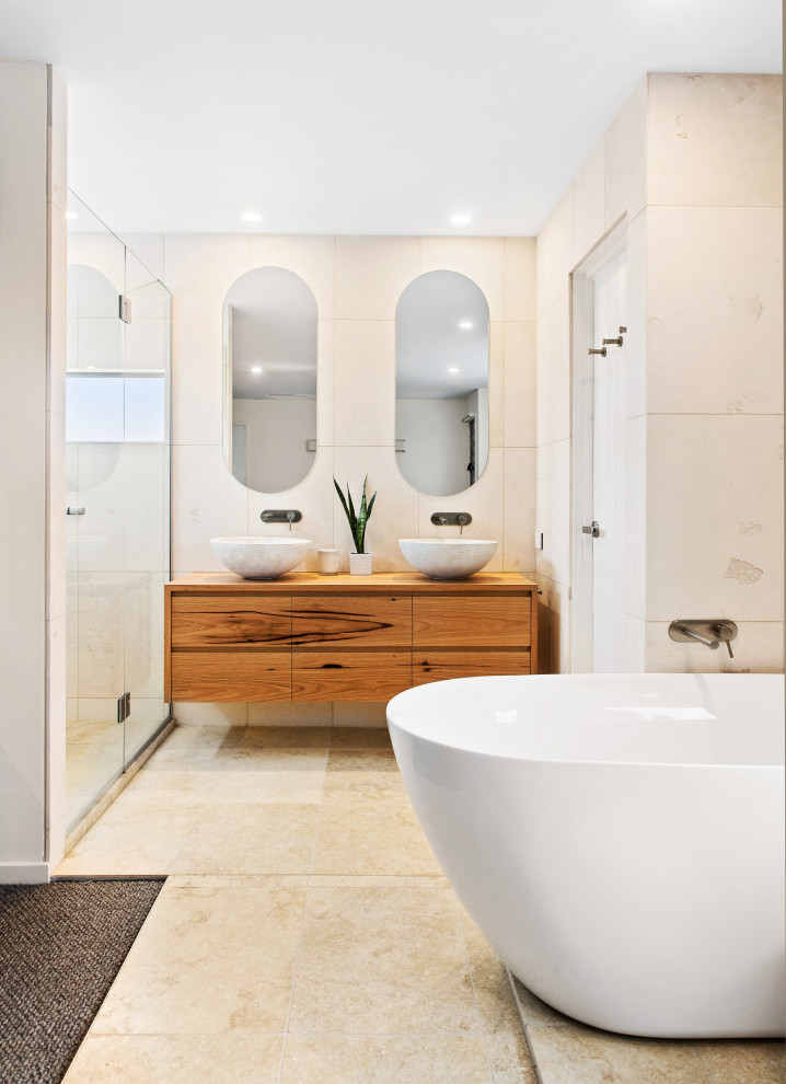 Immagine di una stanza da bagno padronale costiera di medie dimensioni con ante in legno chiaro, piastrelle di pietra calcarea, pavimento in pietra calcarea, toilette, due lavabi e mobile bagno sospeso