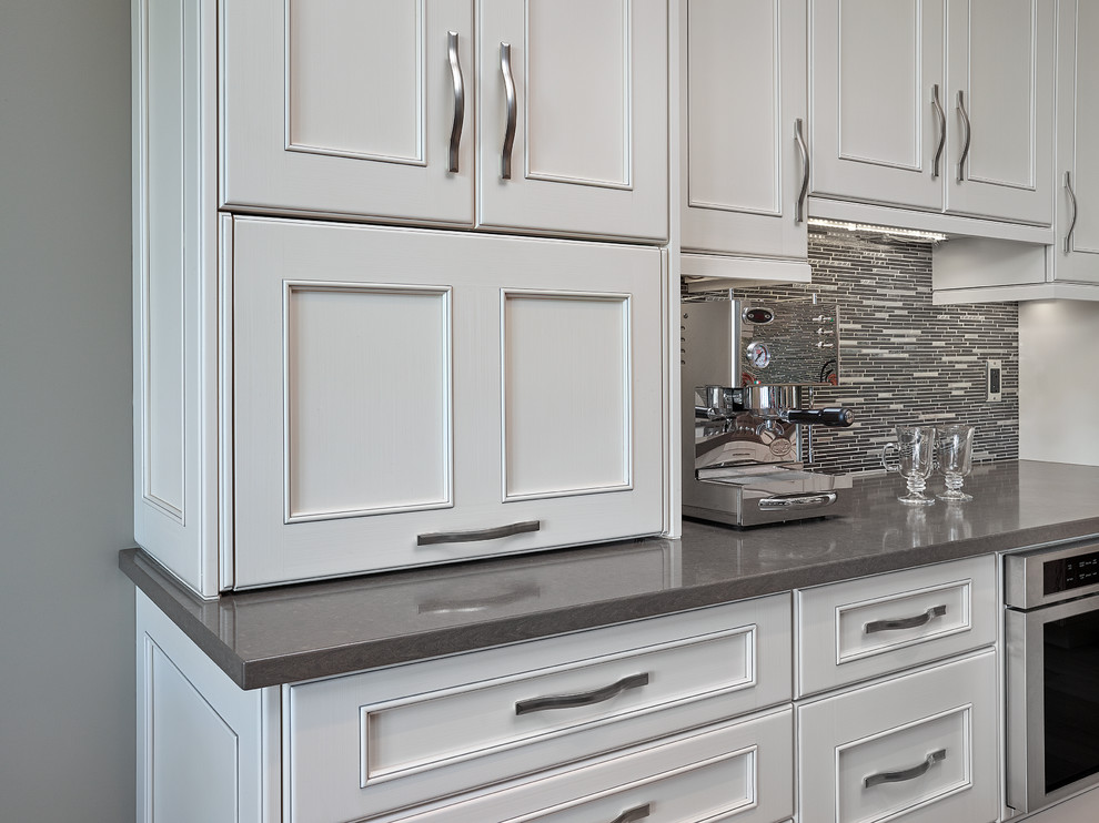 Large transitional l-shaped light wood floor and beige floor eat-in kitchen photo in Edmonton with a farmhouse sink, recessed-panel cabinets, white cabinets, quartz countertops, gray backsplash, matchstick tile backsplash, stainless steel appliances and an island