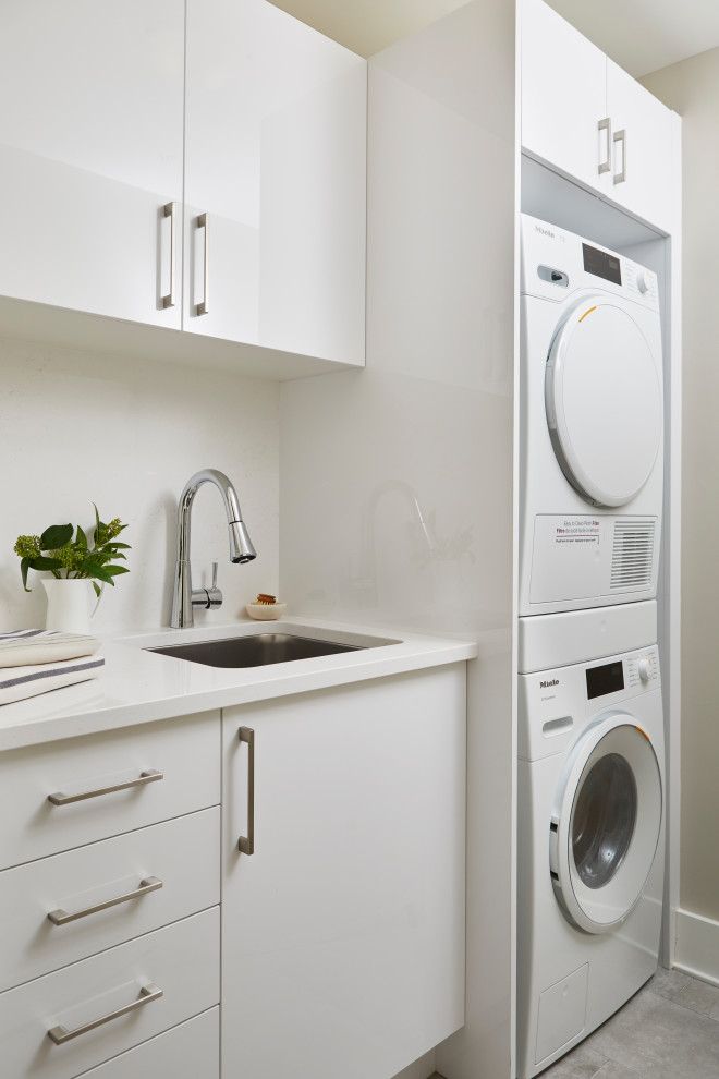 Design ideas for a small contemporary single-wall dedicated laundry room in Toronto with an undermount sink, flat-panel cabinets, white cabinets, quartz benchtops, white splashback, engineered quartz splashback, beige walls, ceramic floors, a stacked washer and dryer, grey floor and white benchtop.