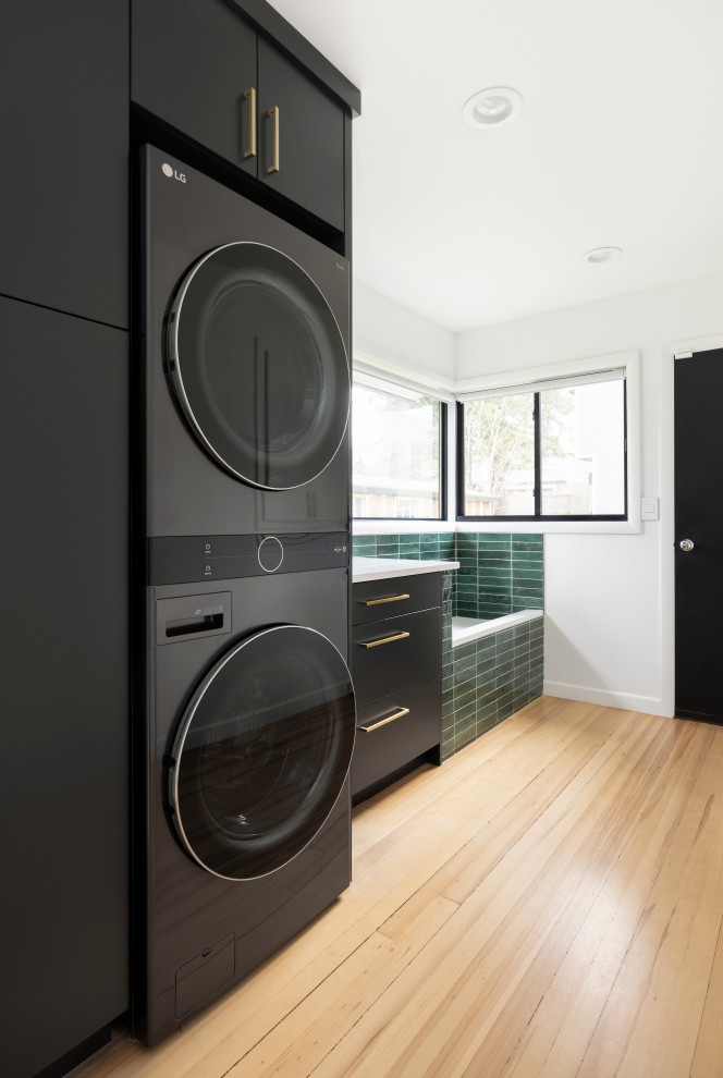 Mid-sized contemporary single-wall utility room in Portland with flat-panel cabinets, black cabinets, quartz benchtops, green splashback, cement tile splashback, white walls, light hardwood floors, a stacked washer and dryer and white benchtop.