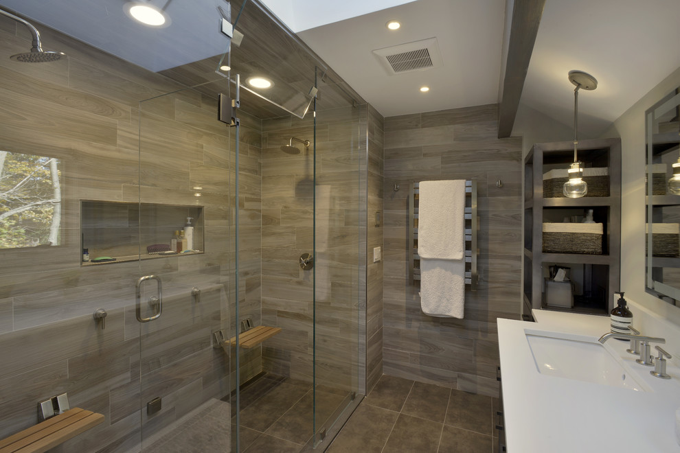 This is an example of a mid-sized modern master bathroom in New York with furniture-like cabinets, grey cabinets, a double shower, a one-piece toilet, brown tile, porcelain tile, white walls, porcelain floors, an undermount sink, glass benchtops, brown floor and a hinged shower door.