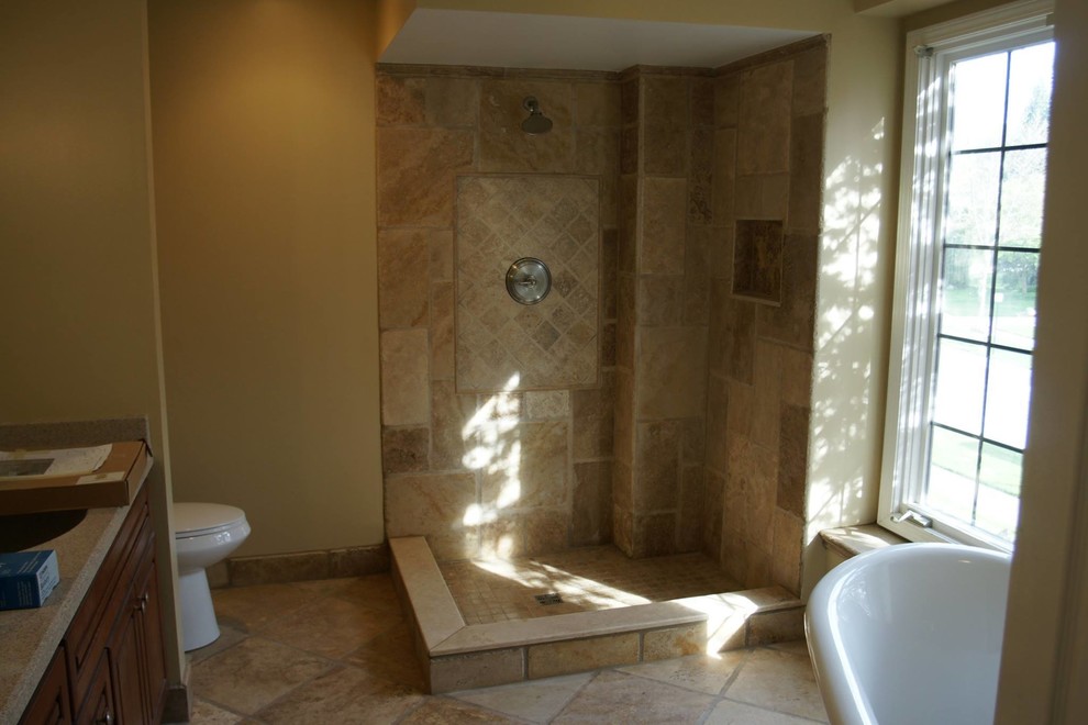 Photo of a mid-sized mediterranean 3/4 bathroom in Detroit with raised-panel cabinets, medium wood cabinets, a freestanding tub, a corner shower, a two-piece toilet, beige tile, limestone, beige walls, limestone floors, an undermount sink, engineered quartz benchtops, beige floor, an open shower and beige benchtops.