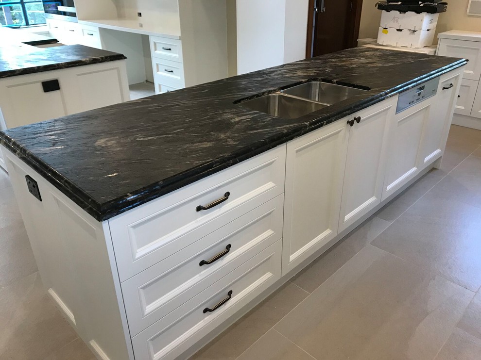 This is an example of an expansive traditional galley open plan kitchen in Sydney with an undermount sink, shaker cabinets, white cabinets, granite benchtops, white splashback, stone slab splashback, black appliances, multiple islands and black benchtop.