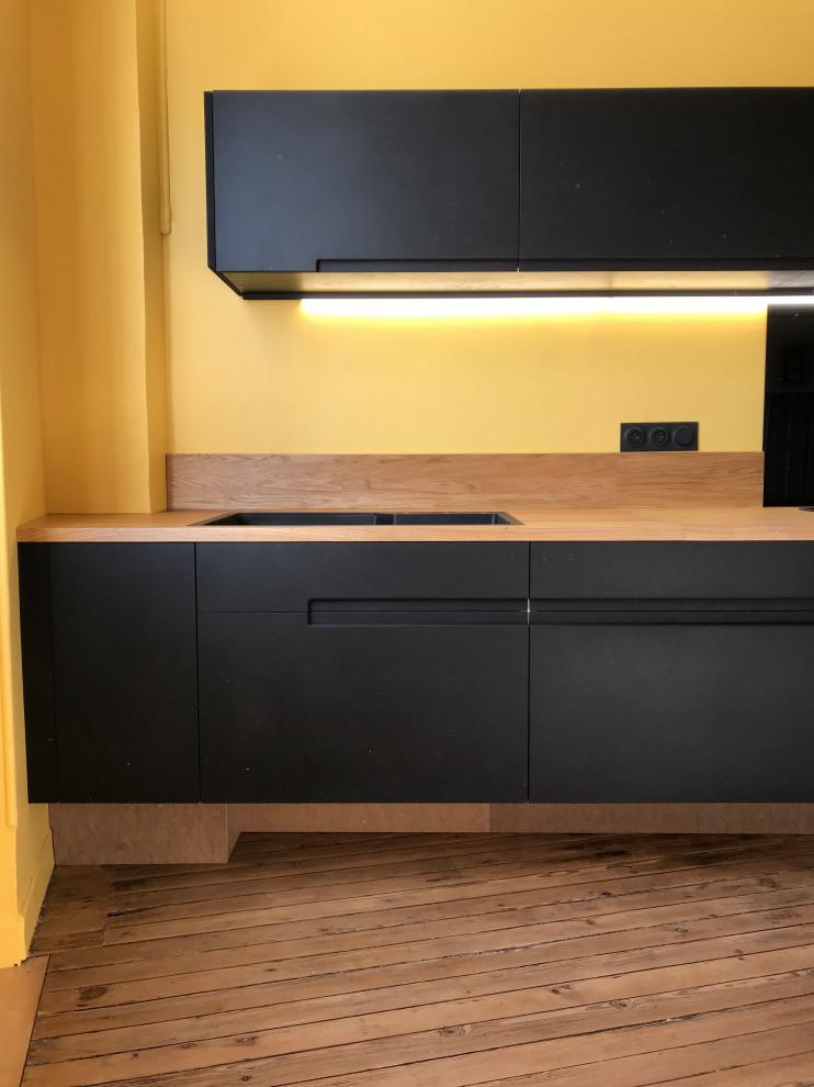 This is an example of a mid-sized contemporary single-wall open plan kitchen in Bordeaux with an undermount sink, flat-panel cabinets, black cabinets, wood benchtops, beige splashback, timber splashback, stainless steel appliances, light hardwood floors, no island, beige floor and beige benchtop.