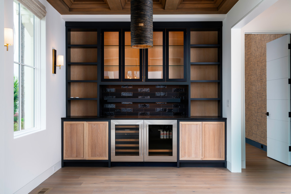 This is an example of a large modern single-wall home bar in Charlotte with no sink, glass-front cabinets, black cabinets, black splashback, ceramic splashback, light hardwood floors, beige floor and black benchtop.