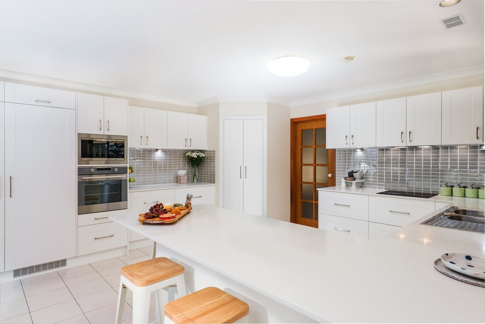 Photo of a large transitional u-shaped eat-in kitchen in Brisbane with a double-bowl sink, shaker cabinets, white cabinets, quartz benchtops, grey splashback, ceramic splashback, stainless steel appliances, ceramic floors and with island.
