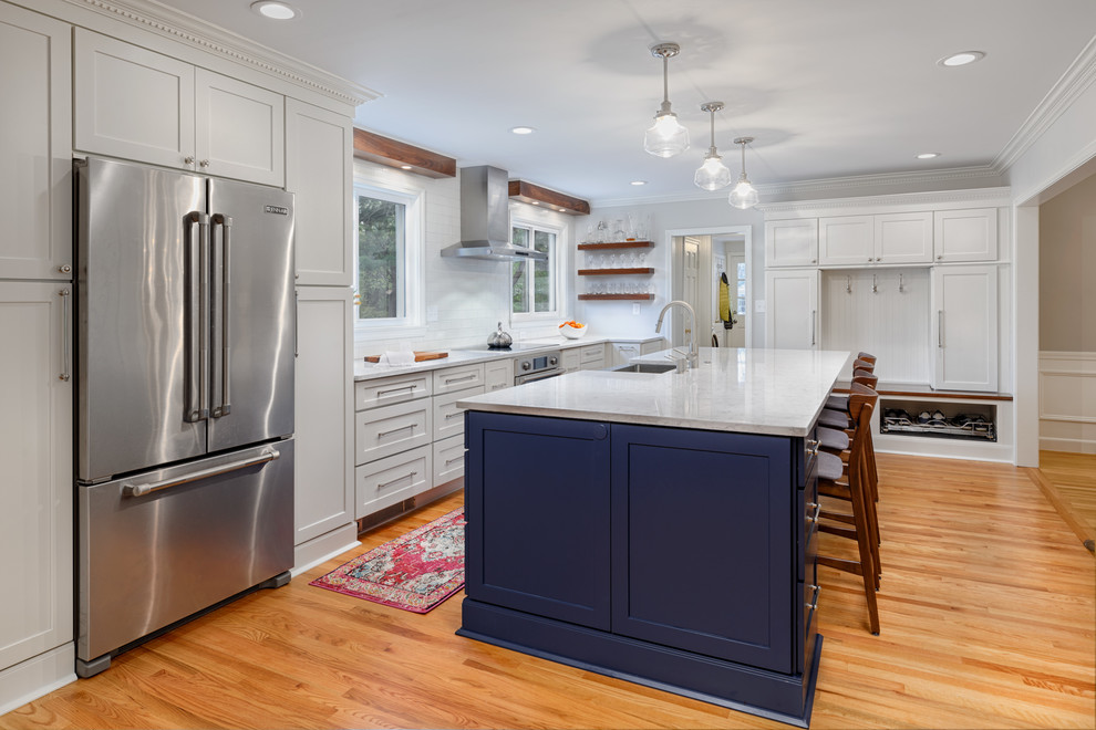 This is an example of an arts and crafts l-shaped open plan kitchen in Boston with a farmhouse sink, shaker cabinets, grey cabinets, white splashback, stainless steel appliances, medium hardwood floors, with island and white benchtop.