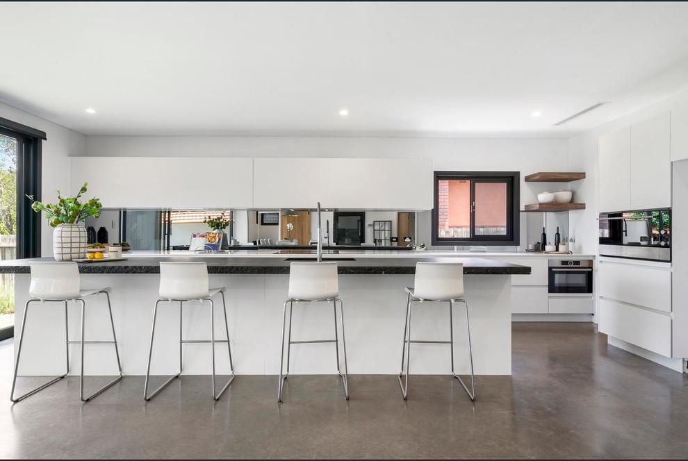 This is an example of a mid-sized contemporary single-wall open plan kitchen in Sydney with an undermount sink, white cabinets, granite benchtops, metallic splashback, mirror splashback, stainless steel appliances, concrete floors, with island and grey floor.