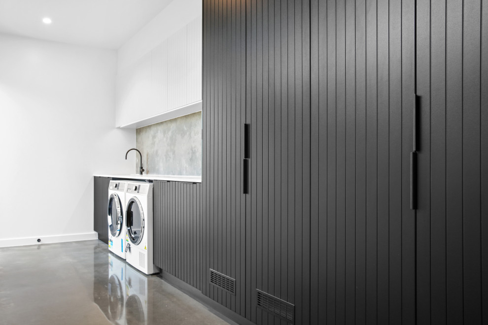 This is an example of a large modern single-wall dedicated laundry room in Other with an undermount sink, beaded inset cabinets, black cabinets, quartz benchtops, grey splashback, stone slab splashback, white walls, concrete floors, a side-by-side washer and dryer, grey floor and white benchtop.