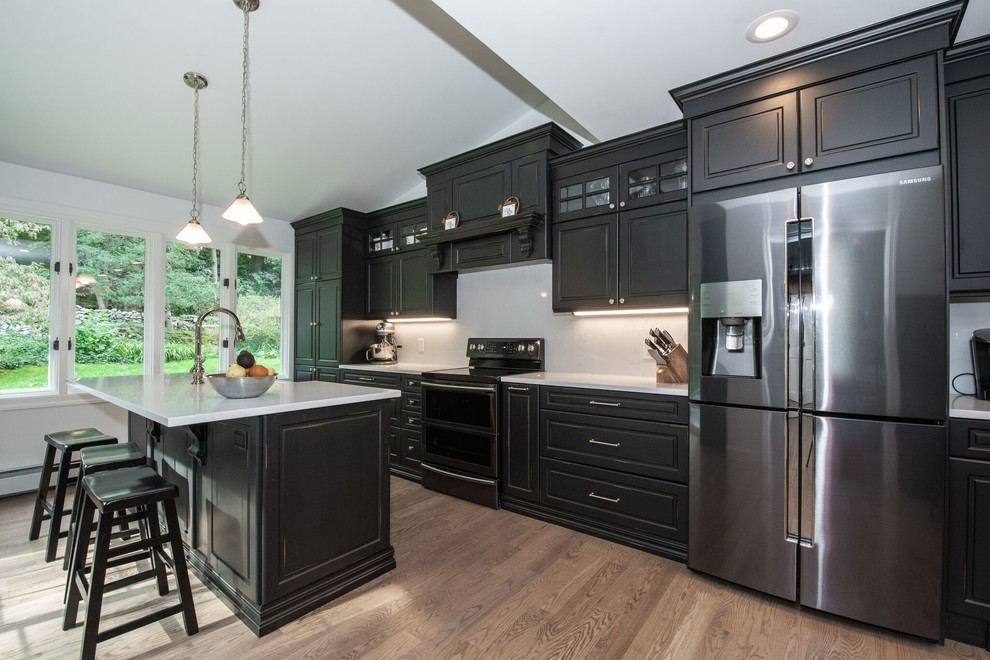 Large traditional single-wall eat-in kitchen in New York with a drop-in sink, raised-panel cabinets, black cabinets, solid surface benchtops, white splashback, glass sheet splashback, stainless steel appliances, medium hardwood floors, with island, brown floor and white benchtop.