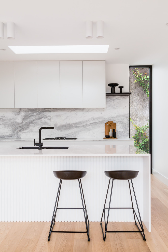 Mid-sized modern galley open plan kitchen in Melbourne with an undermount sink, flat-panel cabinets, white cabinets, solid surface benchtops, grey splashback, marble splashback, black appliances, plywood floors, with island, beige floor, white benchtop and recessed.