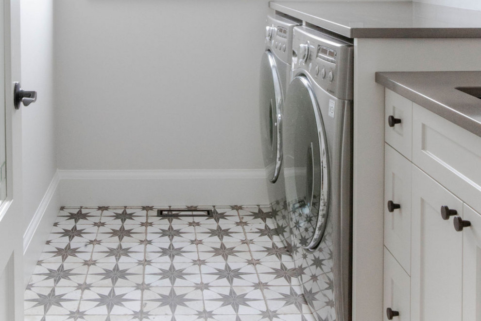 Inredning av en klassisk mellanstor grå parallell grått tvättstuga enbart för tvätt, med en integrerad diskho, släta luckor, vita skåp, bänkskiva i kvarts, vitt stänkskydd, stänkskydd i tunnelbanekakel, vita väggar, en tvättmaskin och torktumlare bredvid varandra och vitt golv