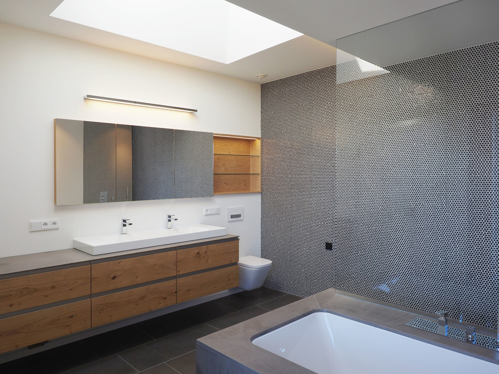 Photo of a mid-sized modern bathroom in Nuremberg with flat-panel cabinets, medium wood cabinets, a curbless shower, a wall-mount toilet, black tile, mosaic tile, white walls, a vessel sink, an undermount tub and marble floors.