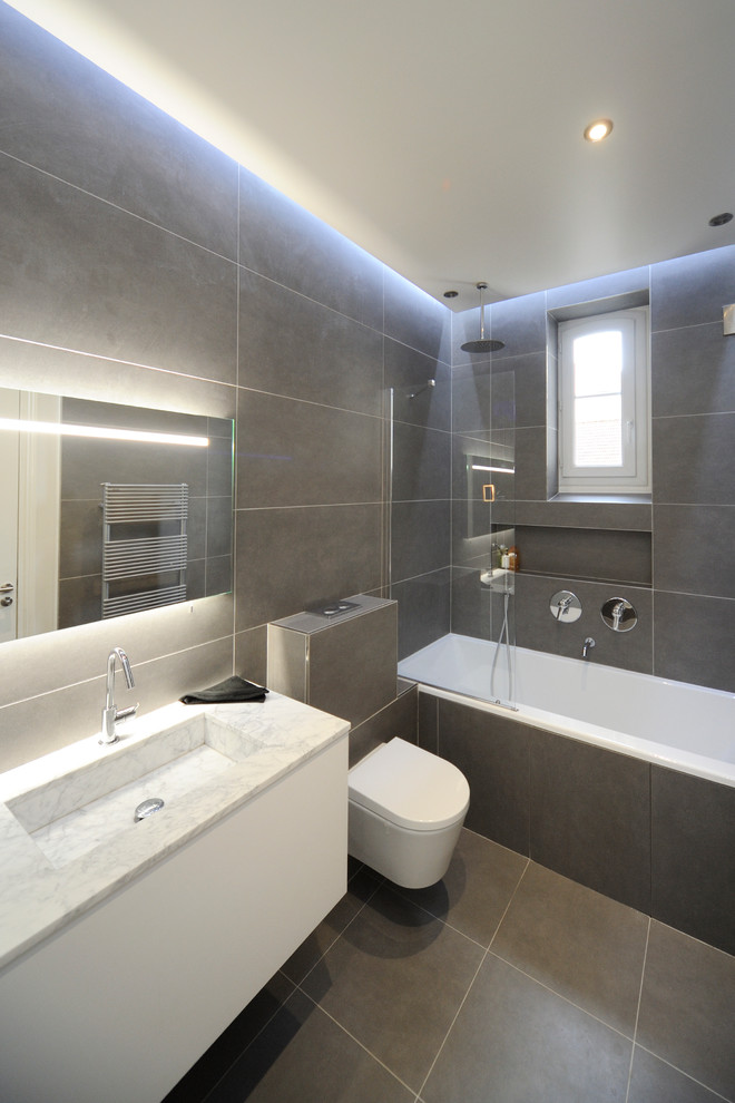 This is an example of a mid-sized contemporary master bathroom in Paris with a drop-in tub, a shower/bathtub combo, a wall-mount toilet, gray tile, grey walls and an integrated sink.