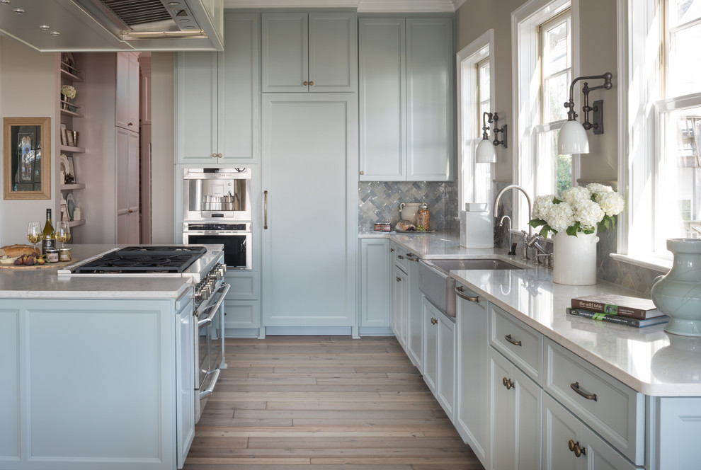 Photo of a large traditional l-shaped kitchen in Milwaukee with a farmhouse sink, blue cabinets, marble benchtops, beige splashback, stainless steel appliances, light hardwood floors and with island.