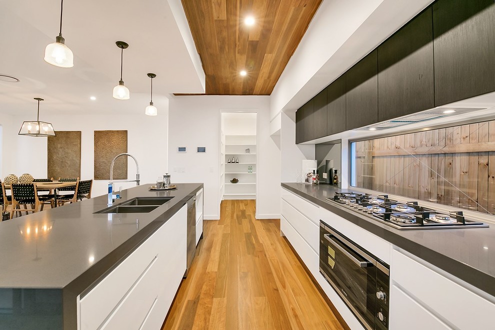Photo of a contemporary galley open plan kitchen in Brisbane with a double-bowl sink, flat-panel cabinets, white cabinets, window splashback, black appliances, medium hardwood floors, with island, brown floor and grey benchtop.