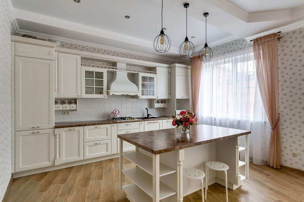 Photo of a traditional single-wall kitchen in Other with a drop-in sink, recessed-panel cabinets, white cabinets, white splashback, with island, brown floor and brown benchtop.
