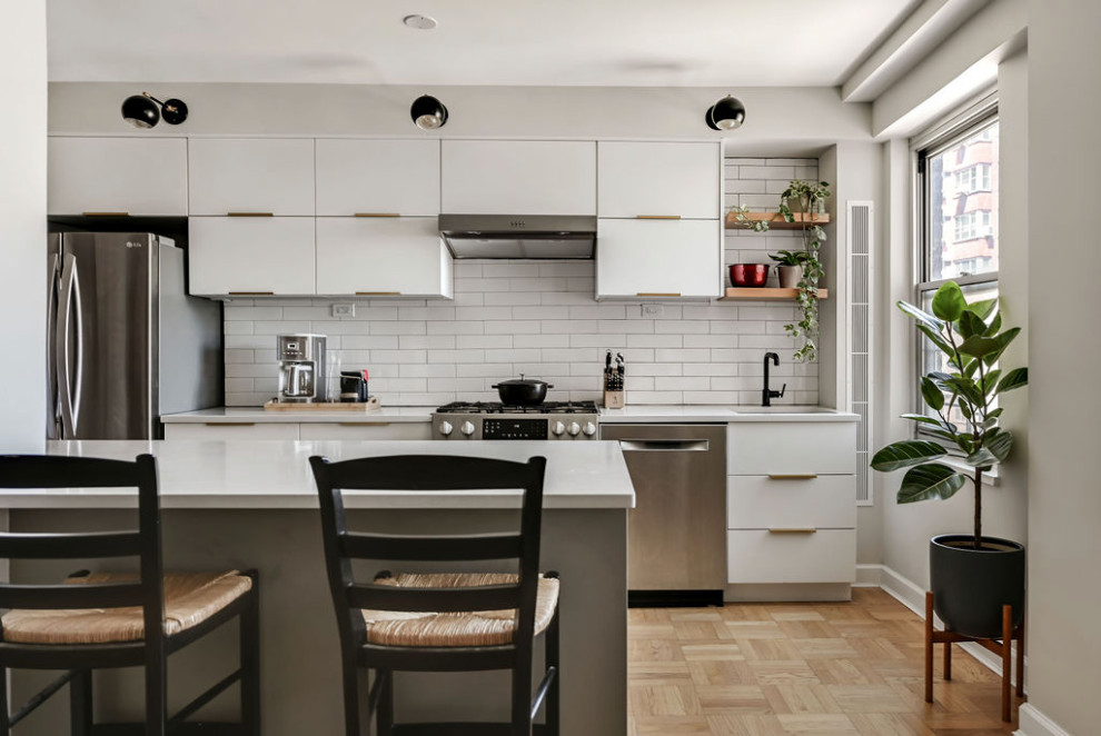 Inspiration for a small transitional galley light wood floor eat-in kitchen remodel in New York with an undermount sink, flat-panel cabinets, white cabinets, quartz countertops, white backsplash, ceramic backsplash, stainless steel appliances, an island and gray countertops