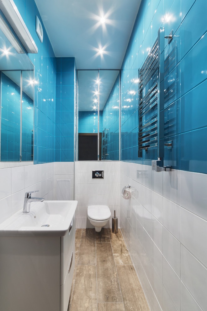 Photo of a mid-sized contemporary powder room in Yekaterinburg with a wall-mount toilet, blue tile, white tile, glass-front cabinets, white cabinets, stone tile, white walls, porcelain floors, a wall-mount sink and brown floor.