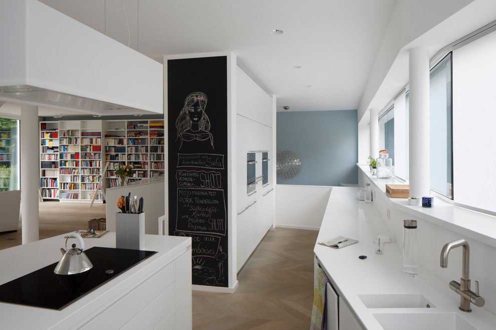 Large contemporary open plan kitchen in Cologne with an integrated sink, flat-panel cabinets, white cabinets, solid surface benchtops, white splashback, light hardwood floors and with island.