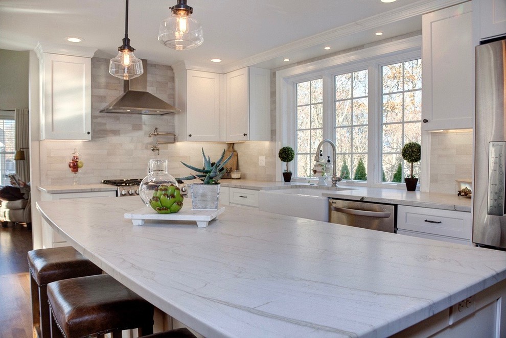 Photo of a mid-sized transitional l-shaped eat-in kitchen in Boston with a farmhouse sink, recessed-panel cabinets, white cabinets, quartzite benchtops, grey splashback, stone tile splashback, stainless steel appliances, medium hardwood floors, with island and brown floor.