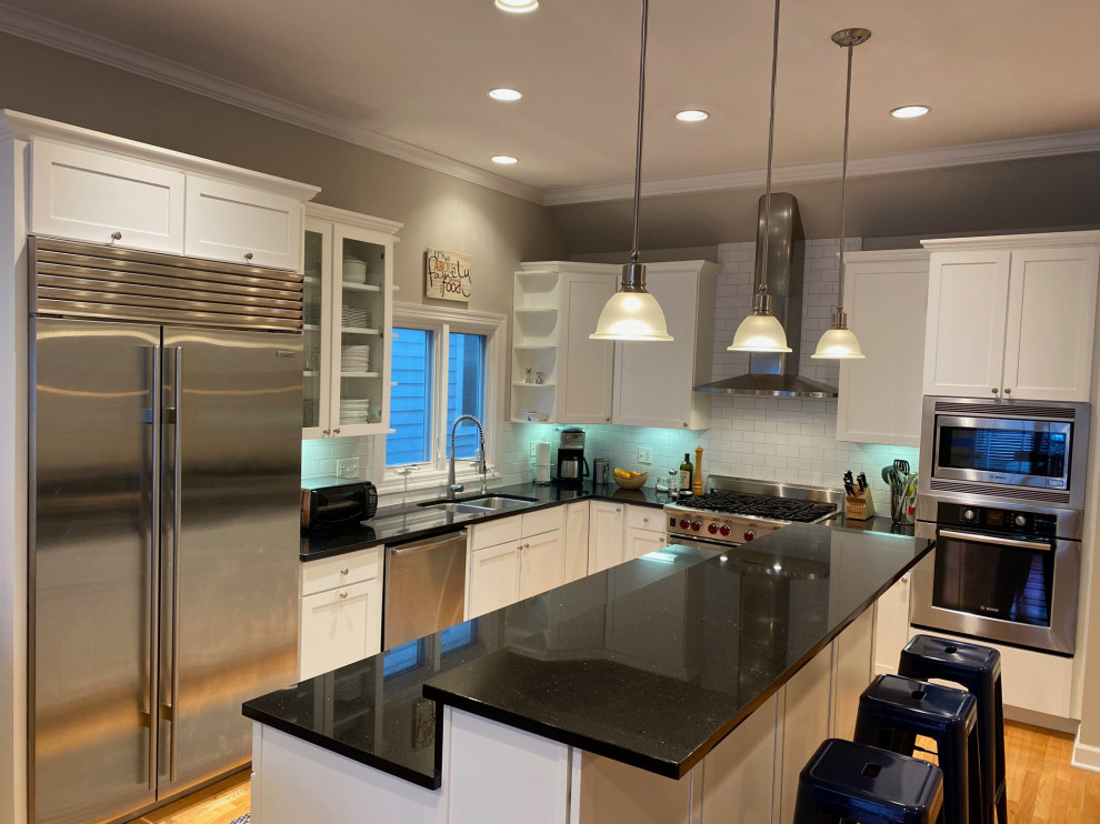 Mid-sized modern l-shaped open plan kitchen in Chicago with a double-bowl sink, shaker cabinets, white cabinets, quartzite benchtops, white splashback, ceramic splashback, stainless steel appliances, light hardwood floors, with island and black benchtop.