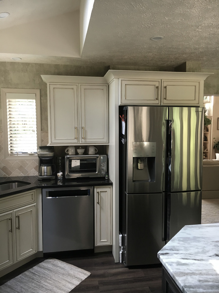 Example of a large transitional u-shaped laminate floor and gray floor eat-in kitchen design with an undermount sink, recessed-panel cabinets, white cabinets, granite countertops, white backsplash, ceramic backsplash, stainless steel appliances, an island and black countertops