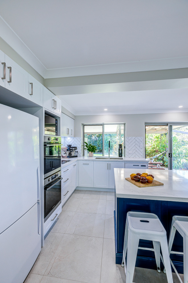 Photo of a large contemporary u-shaped kitchen pantry in Brisbane with a double-bowl sink, shaker cabinets, white cabinets, quartz benchtops, white splashback, subway tile splashback, stainless steel appliances, ceramic floors, with island, beige floor and white benchtop.