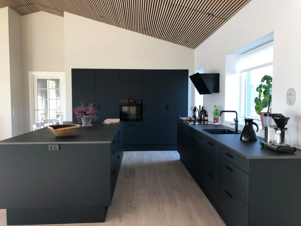 Inspiration for a large contemporary l-shaped kitchen in Aarhus with an integrated sink, flat-panel cabinets, black cabinets, light hardwood floors, with island, beige floor and black benchtop.