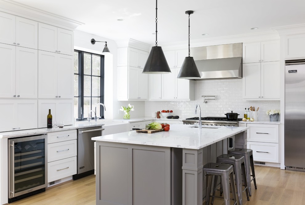 Inspiration for a large country l-shaped kitchen in DC Metro with a farmhouse sink, shaker cabinets, white cabinets, marble benchtops, white splashback, subway tile splashback, stainless steel appliances, with island, white benchtop, medium hardwood floors and brown floor.