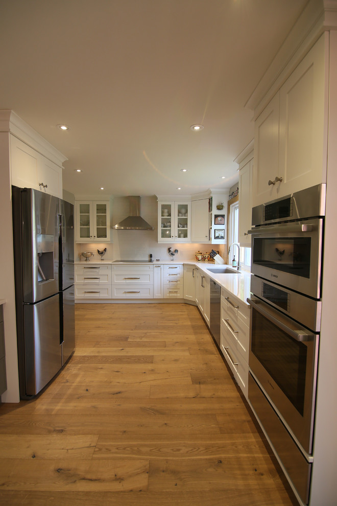 Inspiration for a large transitional l-shaped medium tone wood floor and brown floor eat-in kitchen remodel with an undermount sink, shaker cabinets, white cabinets, stainless steel appliances, an island, solid surface countertops and white backsplash
