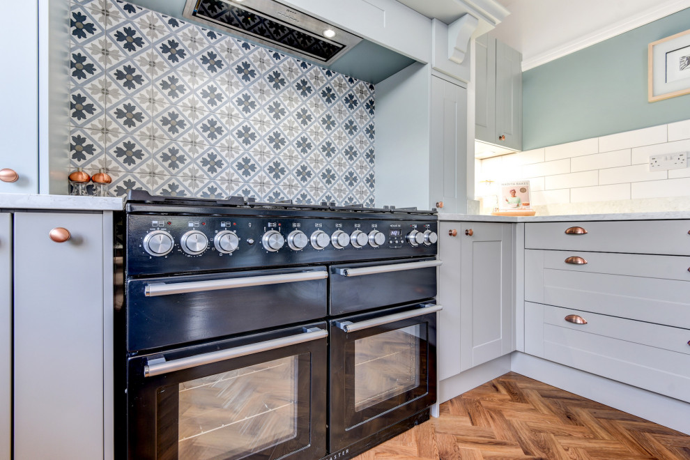 Photo of a large classic grey and teal u-shaped open plan kitchen in Sussex with a belfast sink, shaker cabinets, blue cabinets, quartz worktops, white splashback, metro tiled splashback, black appliances, medium hardwood flooring, a breakfast bar, brown floors, white worktops and a chimney breast.