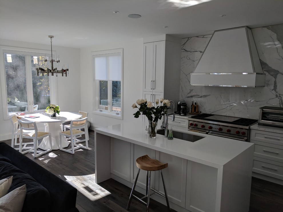 Large contemporary l-shaped open plan kitchen in Toronto with an undermount sink, shaker cabinets, white cabinets, solid surface benchtops, white splashback, marble splashback, panelled appliances, dark hardwood floors, with island and brown floor.