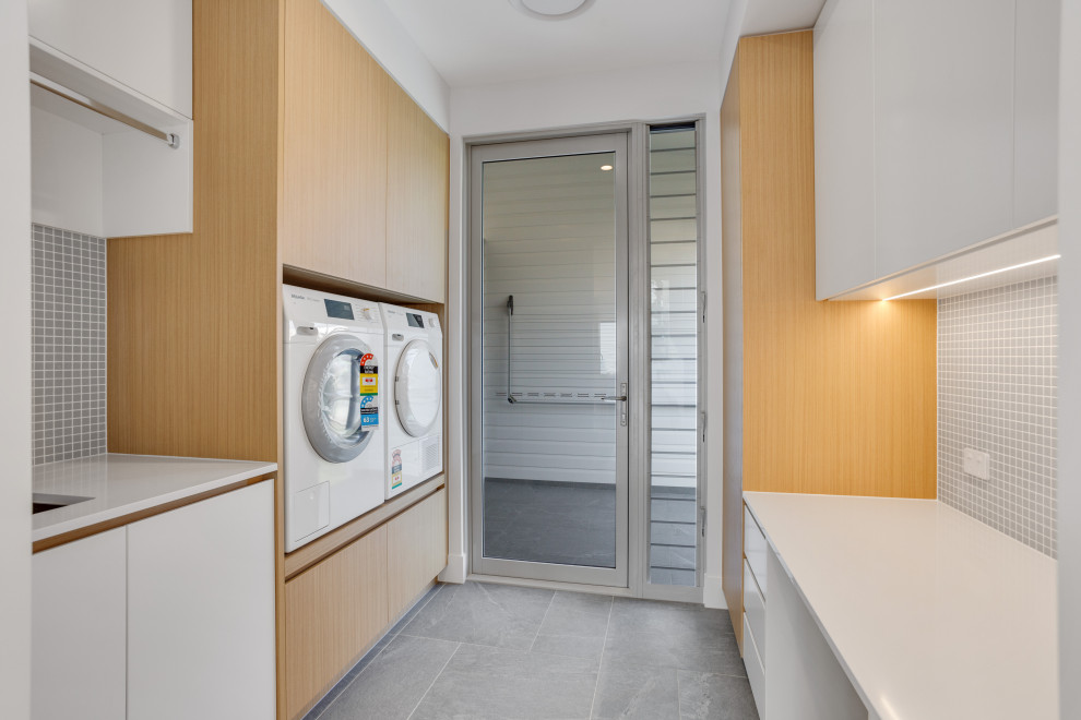Design ideas for a mid-sized beach style galley dedicated laundry room in Brisbane with an undermount sink, brown cabinets, quartz benchtops, grey splashback, mosaic tile splashback, grey walls, porcelain floors, a stacked washer and dryer, grey floor and white benchtop.
