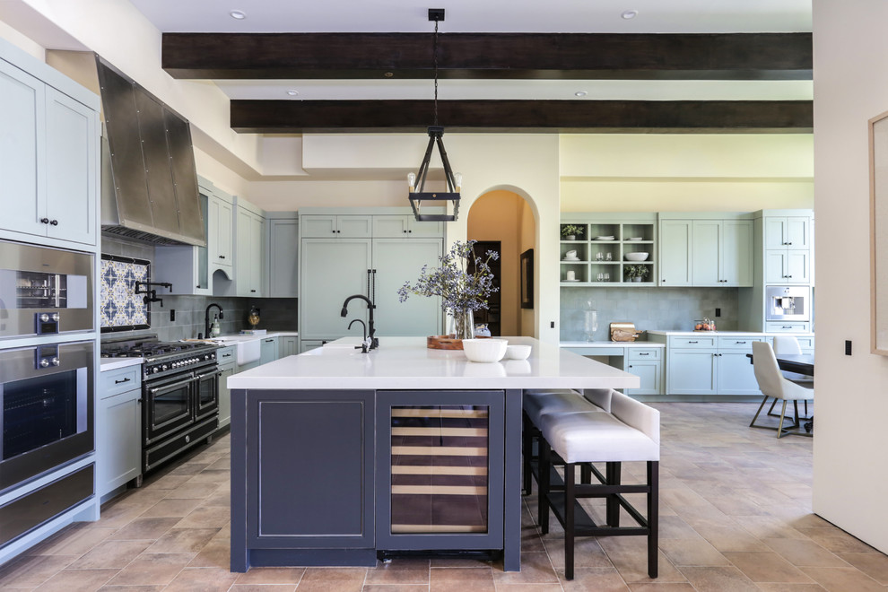 This is an example of a mediterranean l-shaped open plan kitchen in Los Angeles with a farmhouse sink, shaker cabinets, blue cabinets, grey splashback, panelled appliances, with island and brown floor.