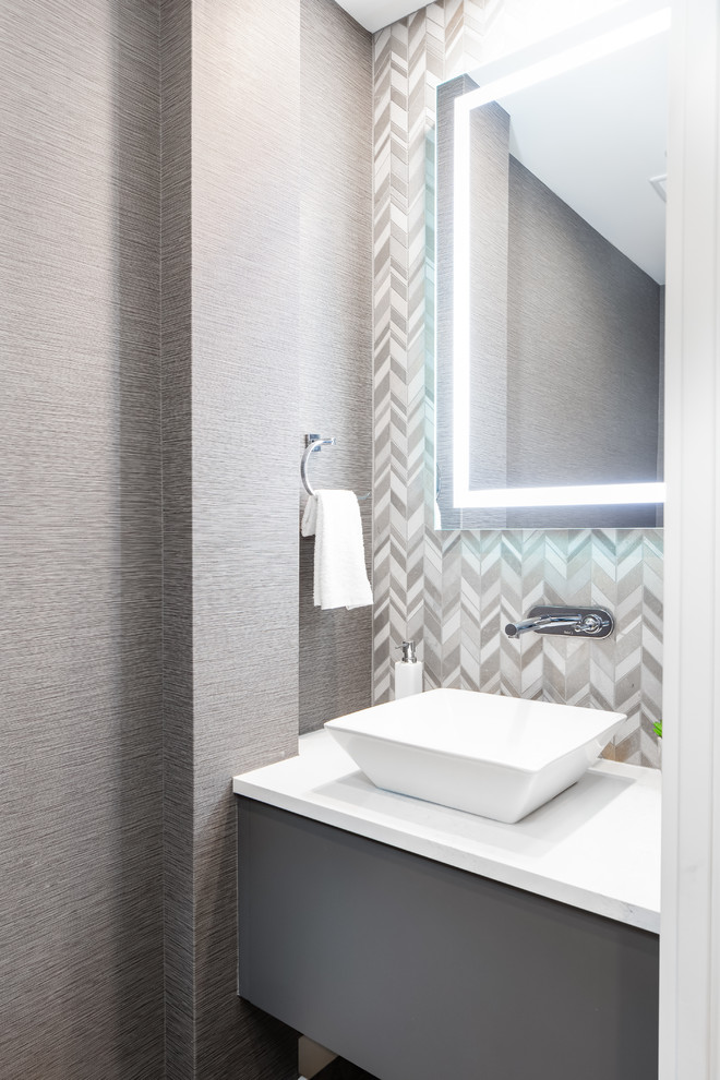 This is an example of a small contemporary powder room in Vancouver with flat-panel cabinets, grey cabinets, multi-coloured tile, mosaic tile, grey walls, medium hardwood floors, a vessel sink, brown floor, white benchtops and engineered quartz benchtops.