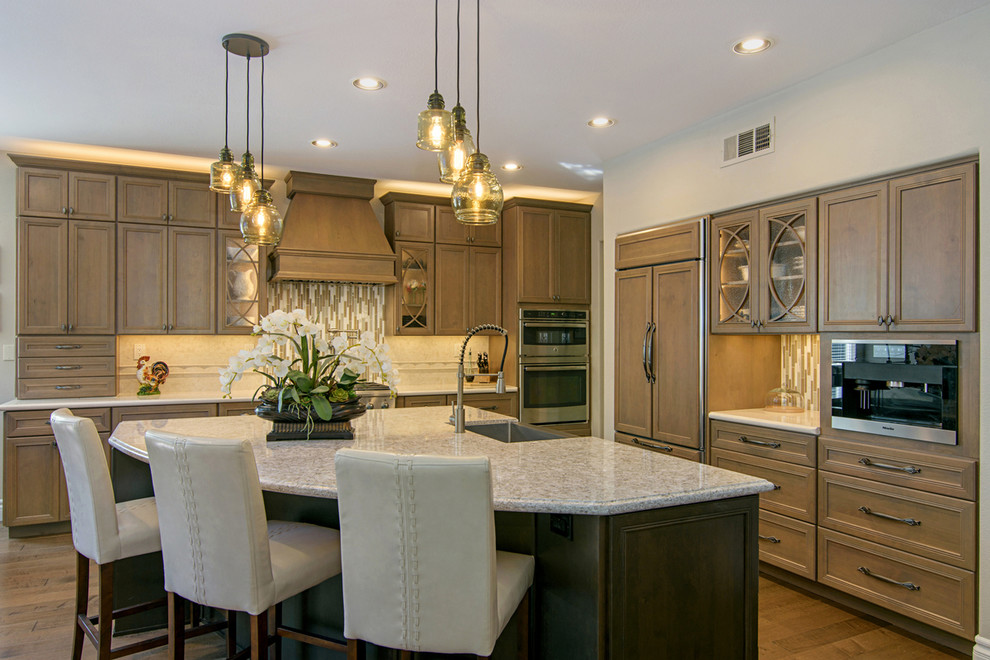 Example of a large transitional l-shaped medium tone wood floor and brown floor open concept kitchen design in San Diego with a farmhouse sink, recessed-panel cabinets, medium tone wood cabinets, beige backsplash, paneled appliances, an island, quartz countertops and travertine backsplash