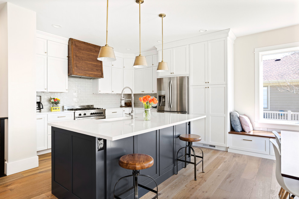 Eat-in kitchen - transitional l-shaped light wood floor eat-in kitchen idea with a farmhouse sink, shaker cabinets, white cabinets, quartz countertops, subway tile backsplash, stainless steel appliances, an island, white countertops and gray backsplash