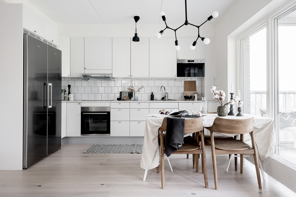 Photo of a mid-sized modern l-shaped open plan kitchen in Gothenburg with a drop-in sink, flat-panel cabinets, white cabinets, laminate benchtops, white splashback, ceramic splashback, stainless steel appliances, light hardwood floors and grey benchtop.