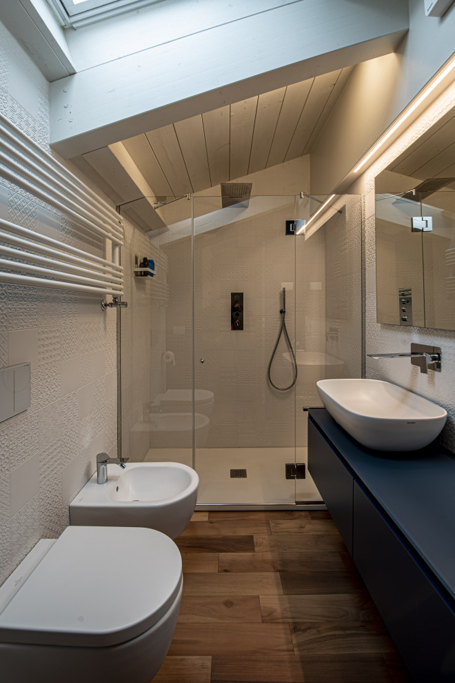 Bild på ett litet blå blått badrum, med släta luckor, blå skåp, en toalettstol med separat cisternkåpa, vit kakel, porslinskakel, vita väggar, målat trägolv, ett fristående handfat, bänkskiva i glas, brunt golv och dusch med gångjärnsdörr