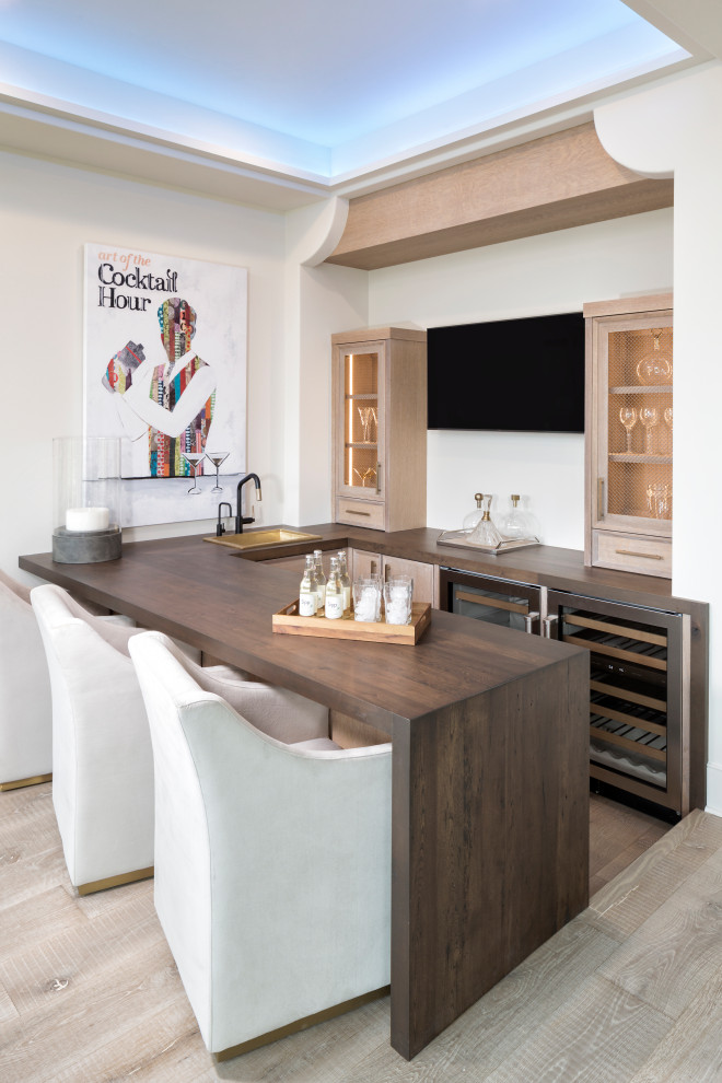 Photo of a transitional u-shaped seated home bar in Minneapolis with a drop-in sink, glass-front cabinets, beige cabinets, wood benchtops, light hardwood floors and brown benchtop.