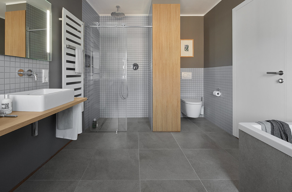 Expansive contemporary bathroom in Bonn with flat-panel cabinets, medium wood cabinets, a drop-in tub, a curbless shower, a wall-mount toilet, gray tile, ceramic tile, grey walls, ceramic floors, a console sink, wood benchtops and brown benchtops.