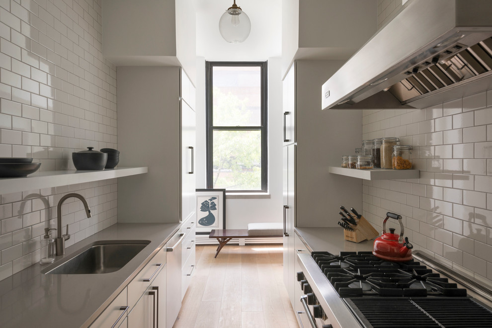 This is an example of a scandinavian galley separate kitchen in New York with an undermount sink, flat-panel cabinets, white cabinets, stainless steel benchtops, white splashback, subway tile splashback, stainless steel appliances, light hardwood floors and no island.