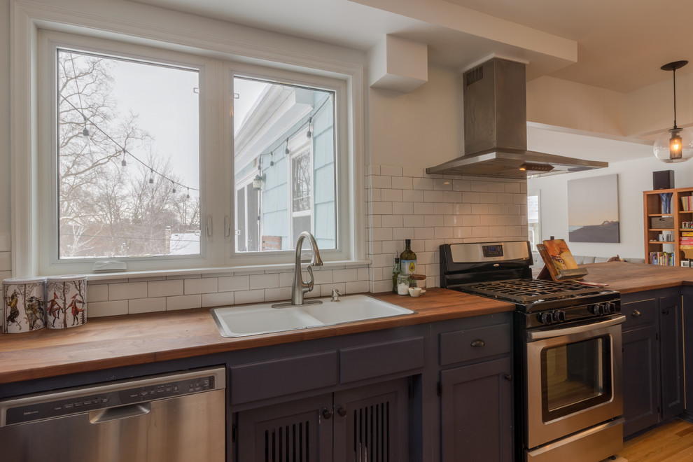 This is an example of a large scandinavian l-shaped open plan kitchen in Minneapolis with a double-bowl sink, recessed-panel cabinets, blue cabinets, wood benchtops, white splashback, subway tile splashback, stainless steel appliances, medium hardwood floors, a peninsula and brown floor.