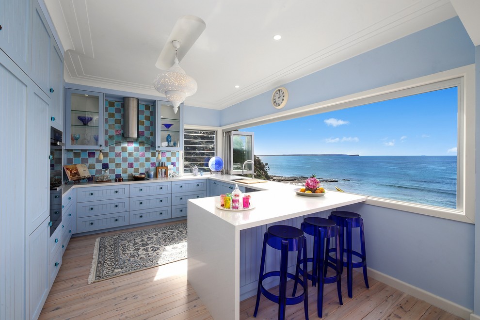 Design ideas for a beach style u-shaped open plan kitchen in Sydney with blue cabinets, multi-coloured splashback, stainless steel appliances, light hardwood floors, a peninsula and shaker cabinets.