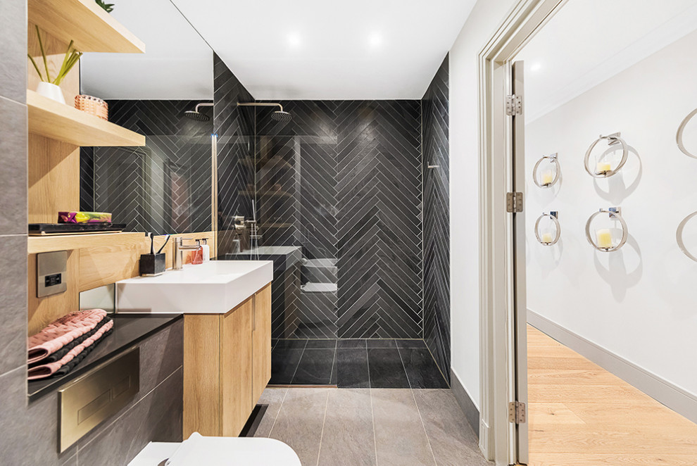 This is an example of a mid-sized contemporary kids bathroom in Essex with flat-panel cabinets, brown cabinets, a freestanding tub, an open shower, a wall-mount toilet, black tile, ceramic tile, multi-coloured walls, ceramic floors, a wall-mount sink, quartzite benchtops, grey floor, an open shower, black benchtops, a single vanity and a floating vanity.