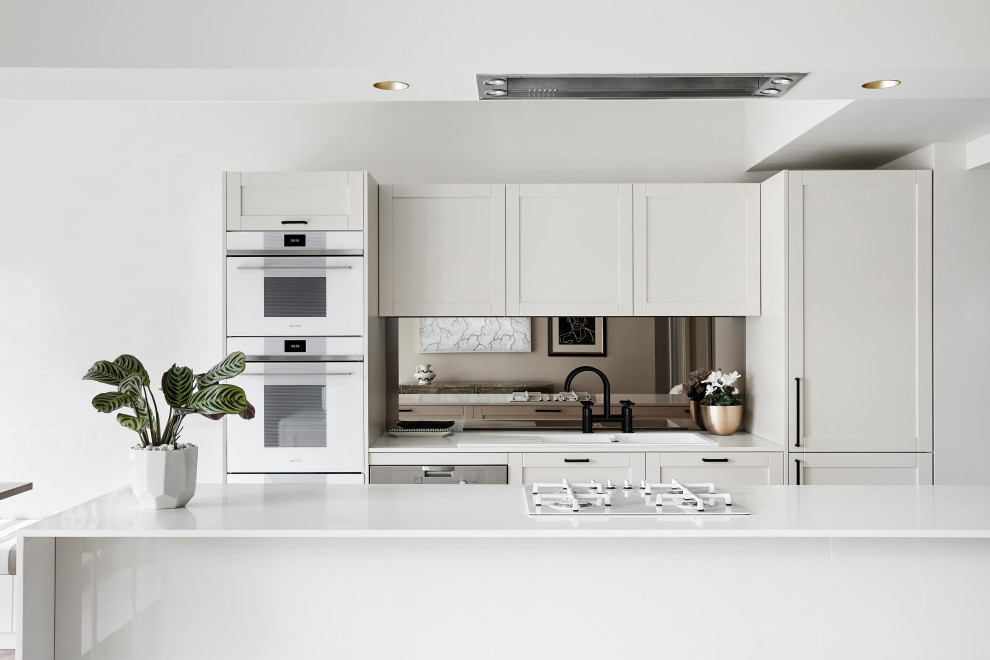 Mid-sized contemporary galley kitchen in Melbourne with a drop-in sink, shaker cabinets, white cabinets, mirror splashback, white appliances, with island and white benchtop.