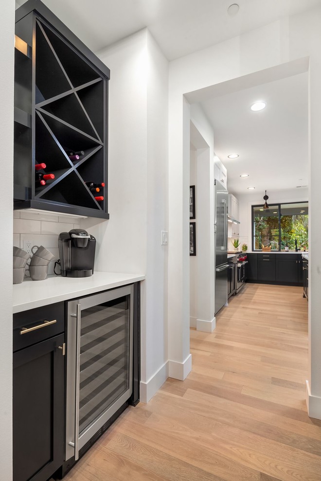 Inspiration for a mid-sized transitional single-wall wet bar in Seattle with shaker cabinets, black cabinets, solid surface benchtops, white splashback, subway tile splashback, light hardwood floors, brown floor and white benchtop.
