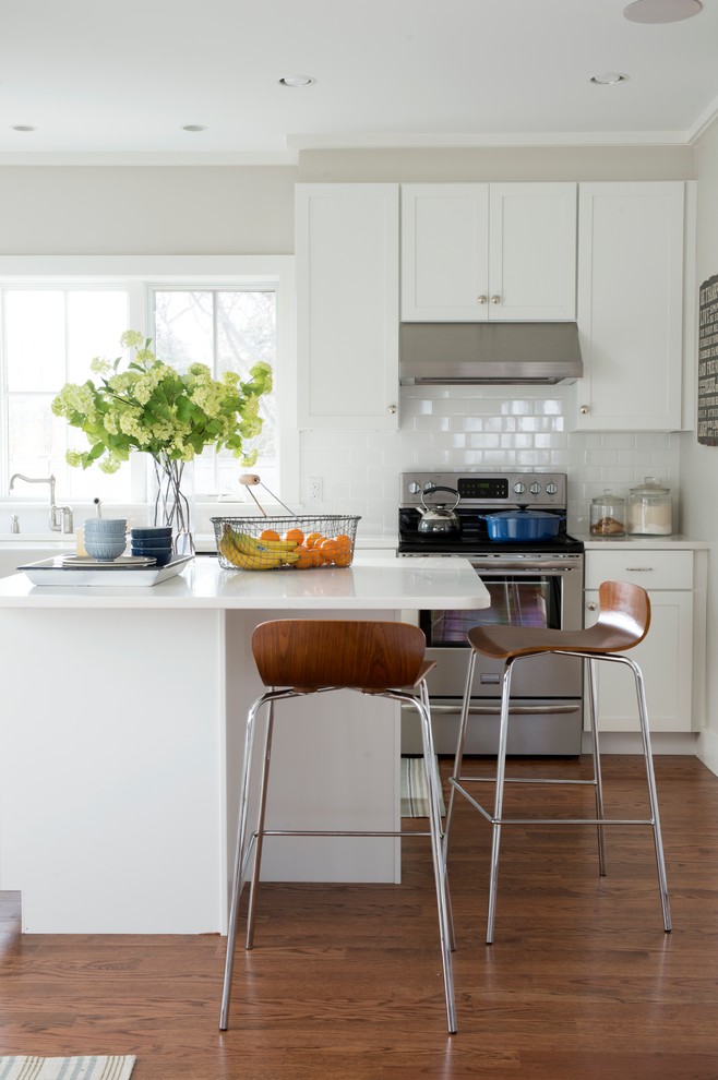 Photo of a mid-sized beach style single-wall eat-in kitchen in New York with shaker cabinets, white cabinets, white splashback, subway tile splashback, stainless steel appliances, medium hardwood floors, with island, an undermount sink, quartz benchtops and brown floor.