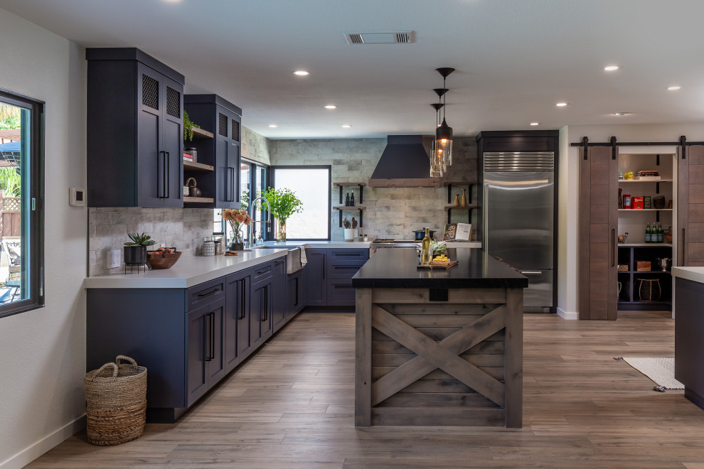 This is an example of a large industrial l-shaped eat-in kitchen in San Francisco with an undermount sink, shaker cabinets, black cabinets, granite benchtops, stone slab splashback, stainless steel appliances, laminate floors, with island, brown floor and black benchtop.
